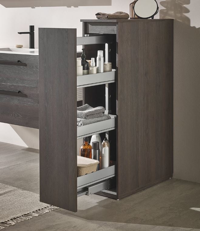 Ein hoher Badezimmerschrank aus dunklem Holz mit ausziehbarem Fach gibt den Blick frei auf drei Regale mit Toilettenartikeln, Flaschen und Handtüchern neben einem modernen Waschbecken.